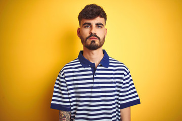 Young man with tattoo wearing striped polo standing over isolated yellow background with serious expression on face. Simple and natural looking at the camera.