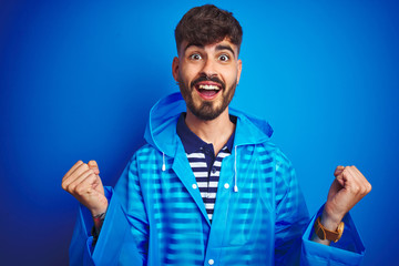 Obraz premium Young handsome man wearing rain coat standing over isolated blue background celebrating surprised and amazed for success with arms raised and open eyes. Winner concept.