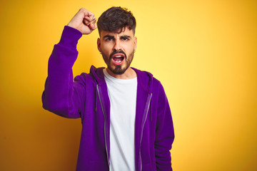 Young man with tattoo wearing sport purple sweatshirt over isolated yellow background angry and mad raising fist frustrated and furious while shouting with anger. Rage and aggressive concept.