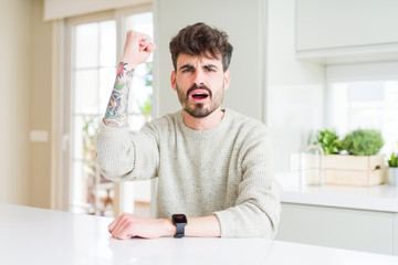 Young man wearing casual sweater sitting on white table angry and mad raising fist frustrated and furious while shouting with anger. Rage and aggressive concept.