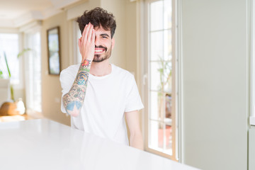 Young man wearing casual t-shirt sitting on white table covering one eye with hand with confident smile on face and surprise emotion.