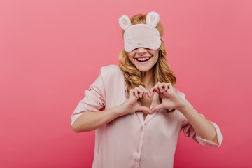 Spectacular curly white woman in eyemask going to sleep. Indoor portrait of positive fair-haired girl posing with pleasure on pink background.