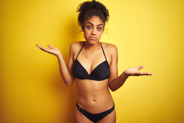 African american woman on vacation wearing bikini standing over isolated yellow background clueless and confused expression with arms and hands raised. Doubt concept.