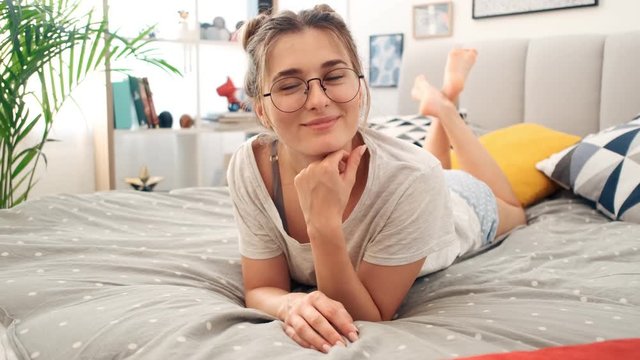 Portrait shot of the young charming Caucasian woman in glasses lying on the bed in the morning in the bedroom and smiling happily to the camera. Close up.