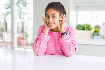 Beautiful african american woman with afro hair wearing casual pink sweater Trying to hear both hands on ear gesture, curious for gossip. Hearing problem, deaf