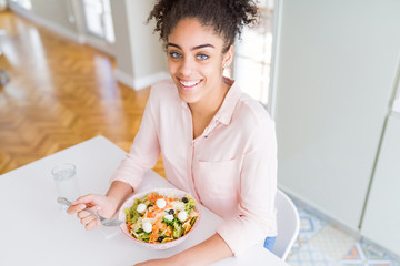 Young african american woman eating healthy pasta salad with a happy face standing and smiling with...