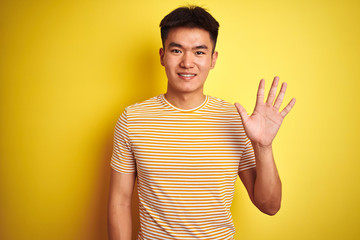 Young asian chinese man wearing t-shirt standing over isolated yellow background showing and pointing up with fingers number five while smiling confident and happy.