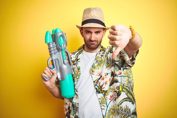 Young man wearing hawaiian flowers shirt holding water gun over yellow isolated background with angry face, negative sign showing dislike with thumbs down, rejection concept