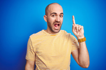 Fototapeta premium Young bald man with beard wearing casual t-shirt over blue isolated background pointing finger up with successful idea. Exited and happy. Number one.