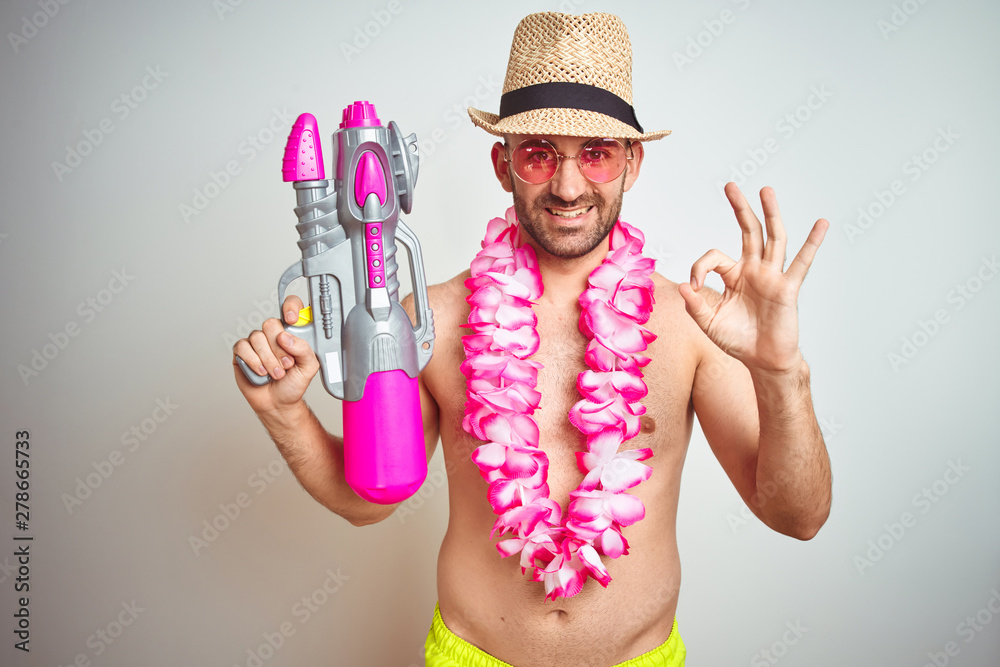 Wall mural Young man wearing summer hat and hawaiian lei flowers holding water gun over isolated background doing ok sign with fingers, excellent symbol