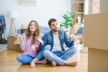 Young beautiful couple moving to a new house sitting on the floor clueless and confused expression with arms and hands raised. Doubt concept.