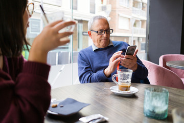 Senior man having a conversation with woman drinking coffee using smartphone and relaxing, chatting at restaurant