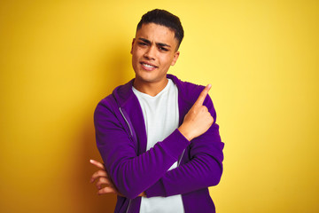 Young brazilian man wearing purple sweatshirt standing over isolated yellow background with a big smile on face, pointing with hand finger to the side looking at the camera.