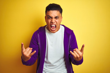 Young brazilian man wearing purple sweatshirt standing over isolated yellow background shouting with crazy expression doing rock symbol with hands up. Music star. Heavy music concept.