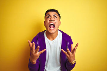 Fototapeta premium Young brazilian man wearing purple sweatshirt standing over isolated yellow background crazy and mad shouting and yelling with aggressive expression and arms raised. Frustration concept.