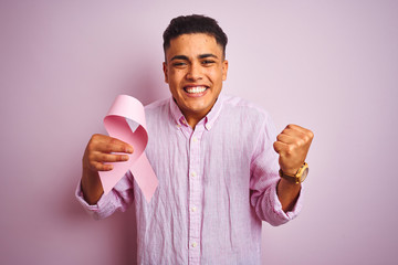Young brazilian man holding cancer ribbon standing over isolated pink background screaming proud...