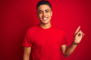 Fototapeta premium Young brazilian man wearing t-shirt standing over isolated red background with a big smile on face, pointing with hand finger to the side looking at the camera.