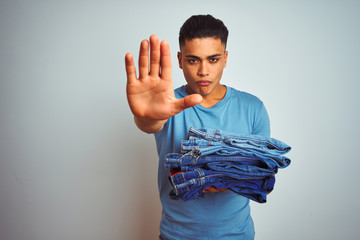 Young brazilian shopkeeper man holding jeans standing over isolated white background with open hand doing stop sign with serious and confident expression, defense gesture