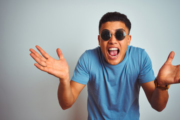 Naklejka premium Young brazilian man wearing blue t-shirt and sunglasses over isolated white background celebrating crazy and amazed for success with arms raised and open eyes screaming excited. Winner concept