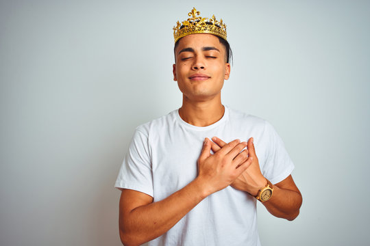 Young brazilian man wearing king crown standing over isolated white background smiling with hands on chest with closed eyes and grateful gesture on face. Health concept.