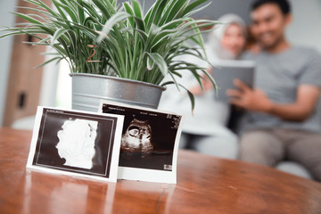 muslim pregnant woman using tablet pc with husband behind ultrasound on table when sitting on the couch in living room