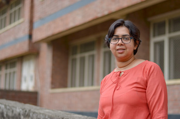 Fototapeta premium Portrait of a confident looking young Indian woman with short hair and spectacles, crossed folded hands in an outdoor setting wearing a traditional North Indian suit dress against a red brick wall