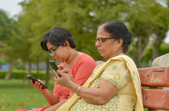 Side Portrait Of Young Indian Woman With An Old North Indian Woman Mother Daughter In Law Looking And Reading On Their Smart Mobile Phones Sitting On A Red Bench In A Park In Delhi, India