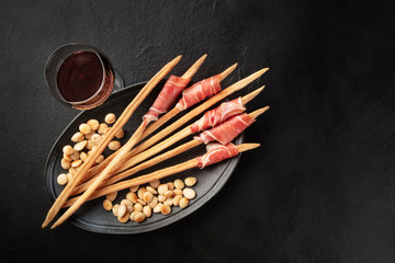 Prosciutto-wrapped Italian grissini with wine and roasted almonds, shot from the top. Italian antipasti with parma ham, overhead photo on a black background with copy space