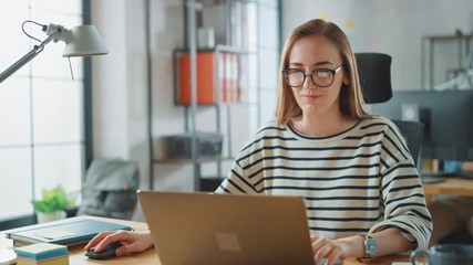 Beautiful Young Woman in Glasses is Working on a Laptop in a Creative Business Agency. They Work in Loft Office. Diverse People Working in the Background. She's in a Good Friendly Mood.