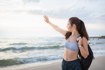 Fototapeta na wymiar Young brunette woman with wireless earbuds, smart watches and black backpack resting after morning workout at the sea shore at sunrise.