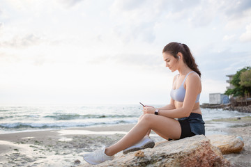 Fototapeta na wymiar Young brunette woman with wireless earbuds and smart watches resting at the sea shore after morning workout at sunrise