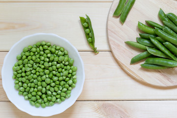 Fresh edible peeled green peas in white plate on wooden table and whole pods on kitchen desk