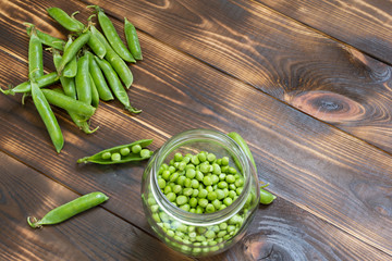 Fresh green peas in glass jar and whole pods on dark wooden table