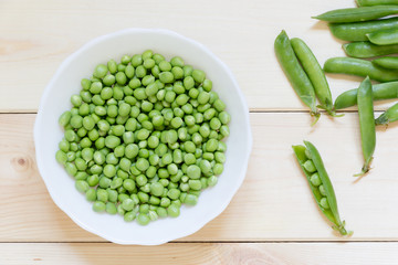 White plate with fresh peeled green peas and whole pods lay on light wooden table