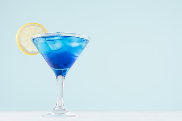 Tropical fresh alcohol cocktail with blue curacao liqueur, ice cube, lemon slice in misted martini glass on soft light white, pastel green background.