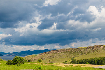 Naklejka premium Blue sky with white clouds, trees, fields and meadows with green grass, against the mountains. Composition of nature. Rural summer landscape.