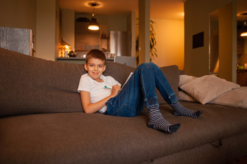 Obraz premium beautiful smiling boy, sitting in couch, looking at camera while writing in his notebook in living room.