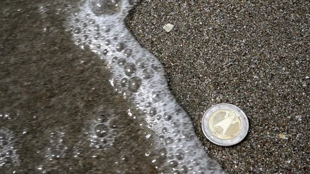 Slow motion sea wave covers euro coin lying on the beach on the sand.