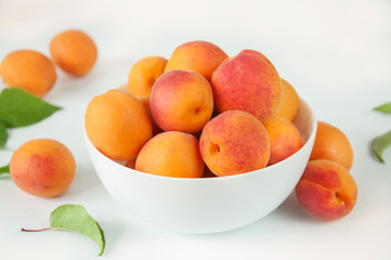 Ripe fruits of apricots with leaves in a white ceramic bowl on a white table.