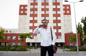 Young handsome business man in sunglasses and white shirt vape e-cigarette on the office building background