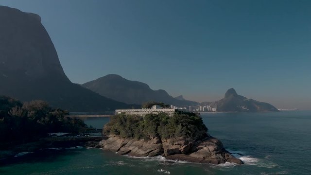 Aerial approach of a small island on the coast of Rio de Janeiro with a construction on top with the well known city peaks of the Two Brothers and Corcovado mountain behind on a sunny hazy afternoon