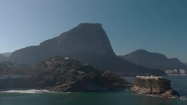 Sideways aerial pan showing the Joatinga beach  on a hazy backlit day with the Gavea mountain behind and an island with recreational construction on top in the foreground