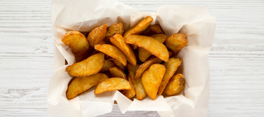 Fried potato wedges in paper box on a white wooden surface. Overhead, top view, flat lay.