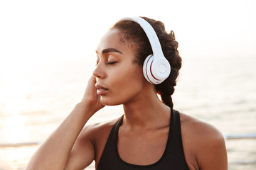 Image of calm pretty woman with headphones standing by seaside in morning