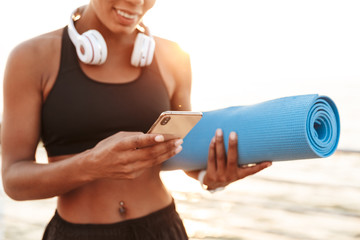 Cropped image of attractive young woman with headphones holding cellphone while standing with fitness mat by seaside in morning