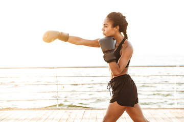 Image of feminine pretty woman training in boxing gloves by seaside in morning