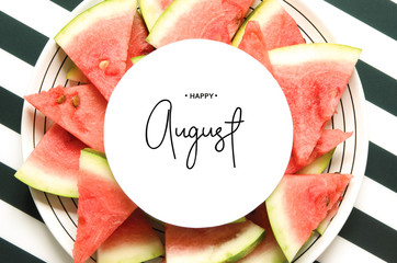 Inscription Happy August. Fresh red watermelon slice  in plate on striped background. Top view, Flat lay. - Image © ireneromanova