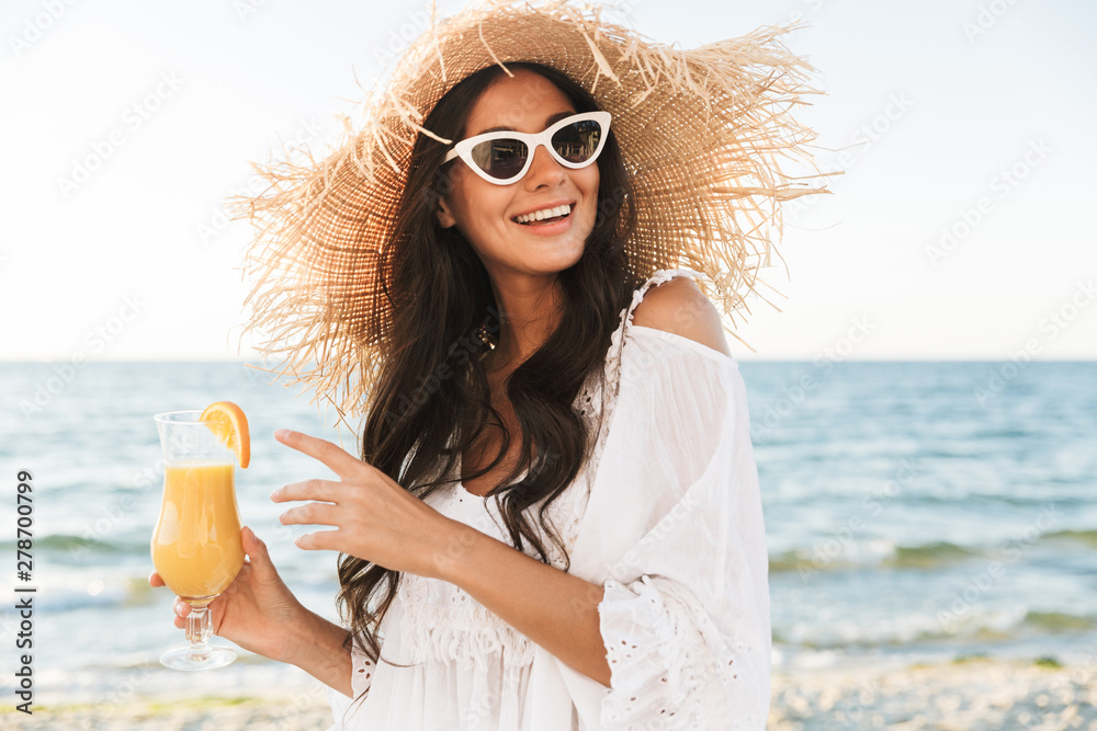 Wall mural photo of happy brunette woman in sunglasses and straw hat walking by seaside with glass of orange ju