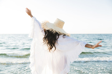 Photo of stylish brunette woman in beach dress and straw hat walking by seaside