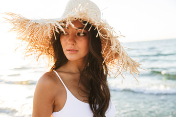 Photo of thoughtful brunette woman in swimsuit and straw hat walking by seaside